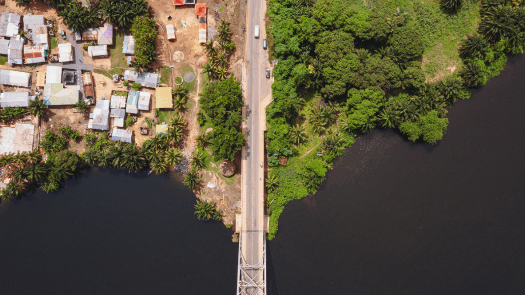 Amazônia_Floresta_Cidade_Foto_Nathaniel Abadji Imagem aérea mostra uma ponte estreita cruzando um rio escuro, ligando duas margens contrastantes — à esquerda, uma área urbana com casas simples e telhados coloridos; à direita, uma região de vegetação densa e verde. A cena evidencia a transição entre ambiente urbano e natureza.