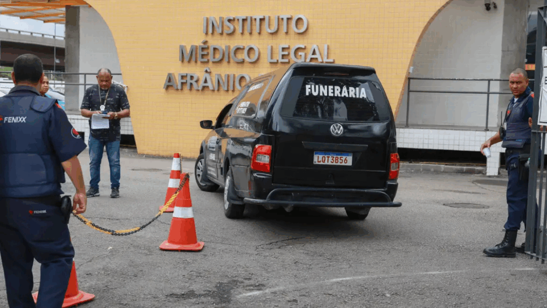 Carro funerário preto com a palavra “FUNERÁRIA” estaciona em frente ao prédio do Instituto Médico Legal Afrânio Peixoto, no Rio de Janeiro. Três homens, incluindo um policial e um agente de segurança, estão próximos ao veículo, enquanto cones laranja delimitam a área de acesso