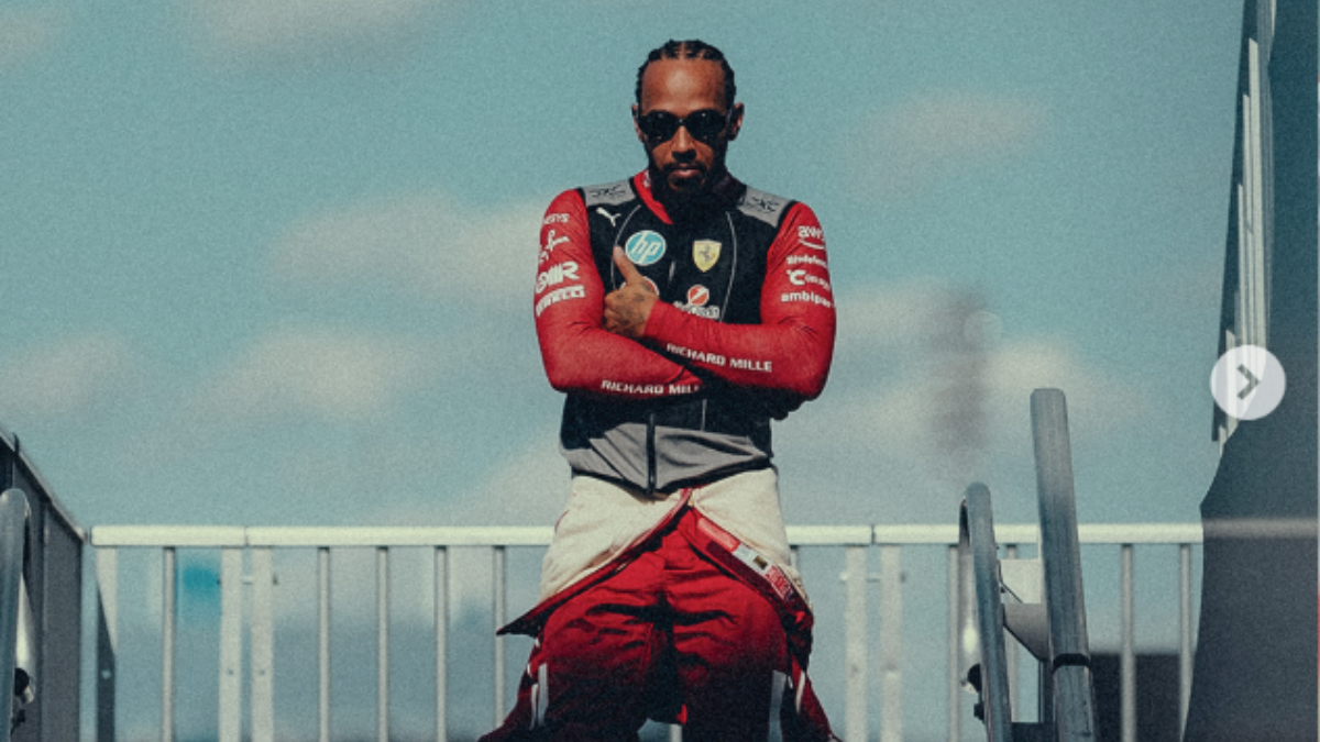 Lewis Hamilton_Ferrari_Foto_Reprodução_Instagram Homem com tranças curtas e óculos escuros posa de braços cruzados, vestindo um macacão de corrida vermelho parcialmente aberto e colete com o logotipo da Ferrari. Ele está em um ambiente externo, sob um céu azul com nuvens suaves, entre estruturas metálicas que lembram o paddock de um autódromo. A iluminação destaca seu semblante confiante e postura firme.