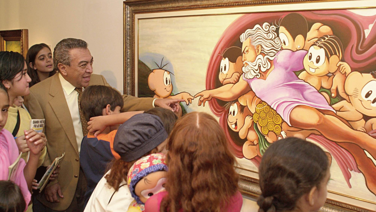 Maurício de Souza_2003_Turma da Mônica_Foto_Agência Brasil Homem de terno bege e gravata clara, cercado por várias crianças, observa e interage com uma pintura colorida emoldurada. A obra é uma releitura bem-humorada da criação de Adão, de Michelangelo, com personagens infantis de desenho animado no lugar das figuras originais. Algumas crianças apontam para o quadro e uma delas segura um boneco. A cena transmite um ambiente educativo e alegre em uma exposição de arte.