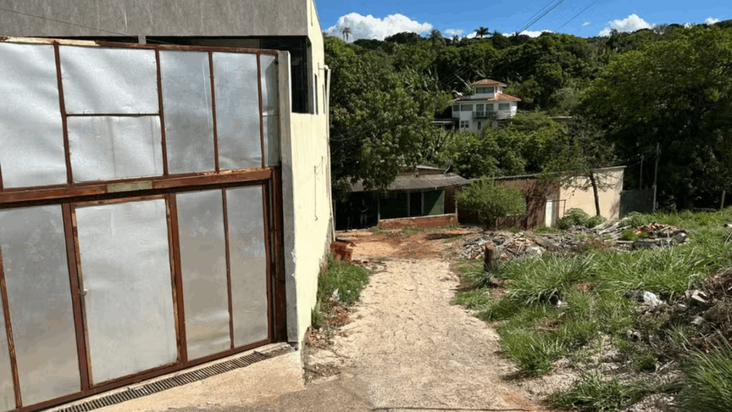 Entrada de uma casa simples com portão metálico em uma rua de terra, cercada por vegetação e outras residências ao fundo, em área residencial de periferia.