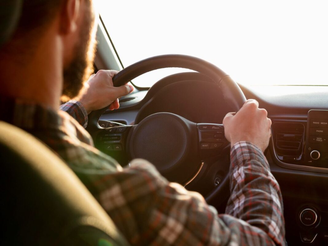 cnh-sem-autoescola-mais-barata A imagem mostra um homem de costas dirigindo com uma camisa xadrez e as duas mãos no volante