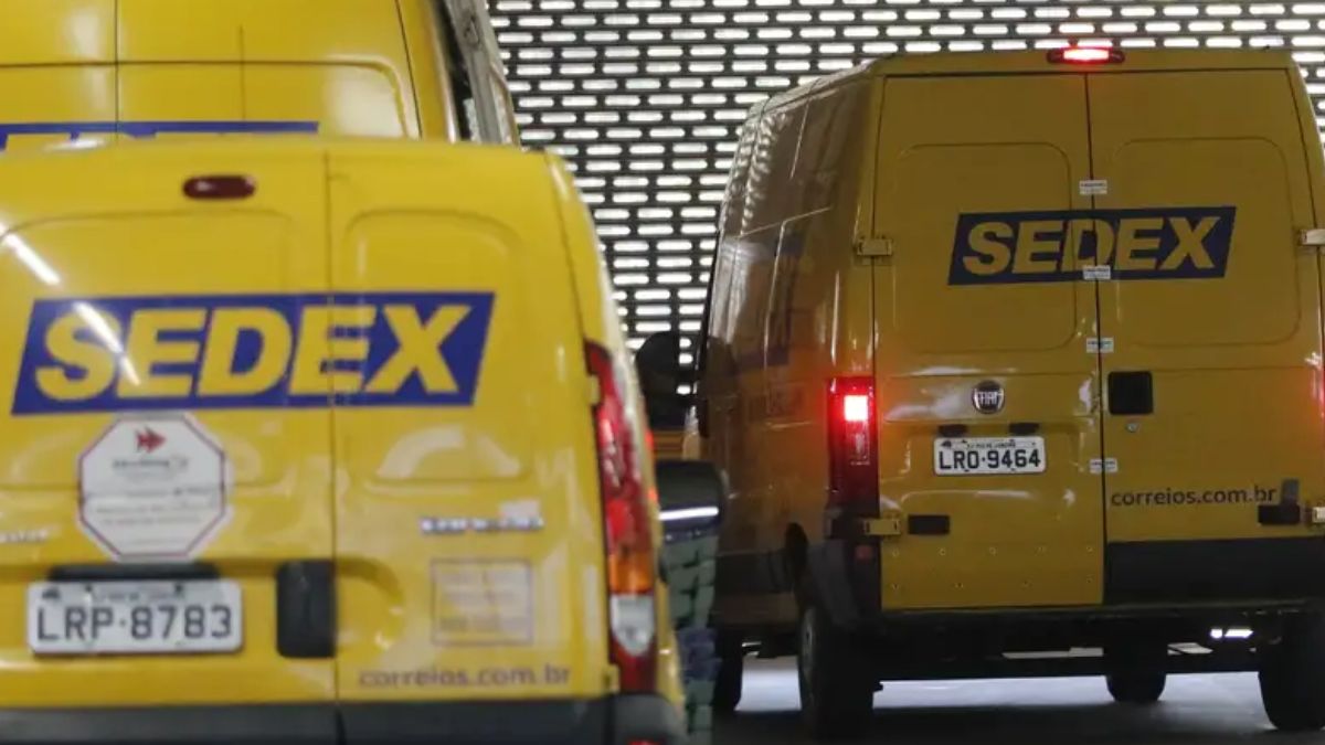 Viaturas do sedex Imagem mostra a traseira de duas vans de entrega dos Correios, ambas na cor amarela característica, em uma área de depósito ou doca de carregamento com uma parede de metal perfurada ao fundo.