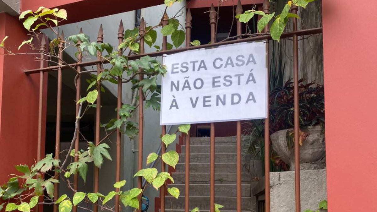 Verticalização em São Paulo Uma foto de um portão de ferro, coberto por algumas trepadeiras de folhas verdes, que revela a entrada de uma casa. O portão está pintado de marrom, e a parede, ao seu lado direito, é de um tom de vermelho terracota. Pendurado no portão, no centro da imagem, há um cartaz retangular branco com letras pretas em caixa alta, que diz: