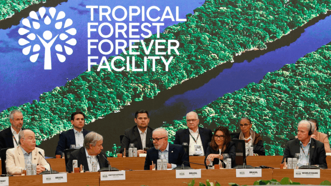 Autoridades e representantes de diferentes países e organizações sentados à mesa durante evento do “Tropical Forest Forever Facility”, com imagem aérea de floresta tropical e rios projetada ao fundo.