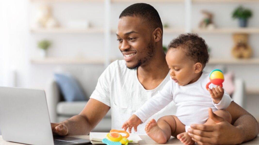 Homem negro sentando de frente para um notebook, com um bebê ao lado